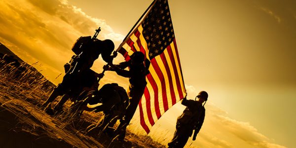 American Soldiers dressed in authentic WWII uniforms stand on a hill in the desert. They are placing the American flag into the ground. Four of the soldiers are assisting each other in standing the flag while the fifth soldier holds the flag up. There is a golden sunset behind them making the men silhouettes.